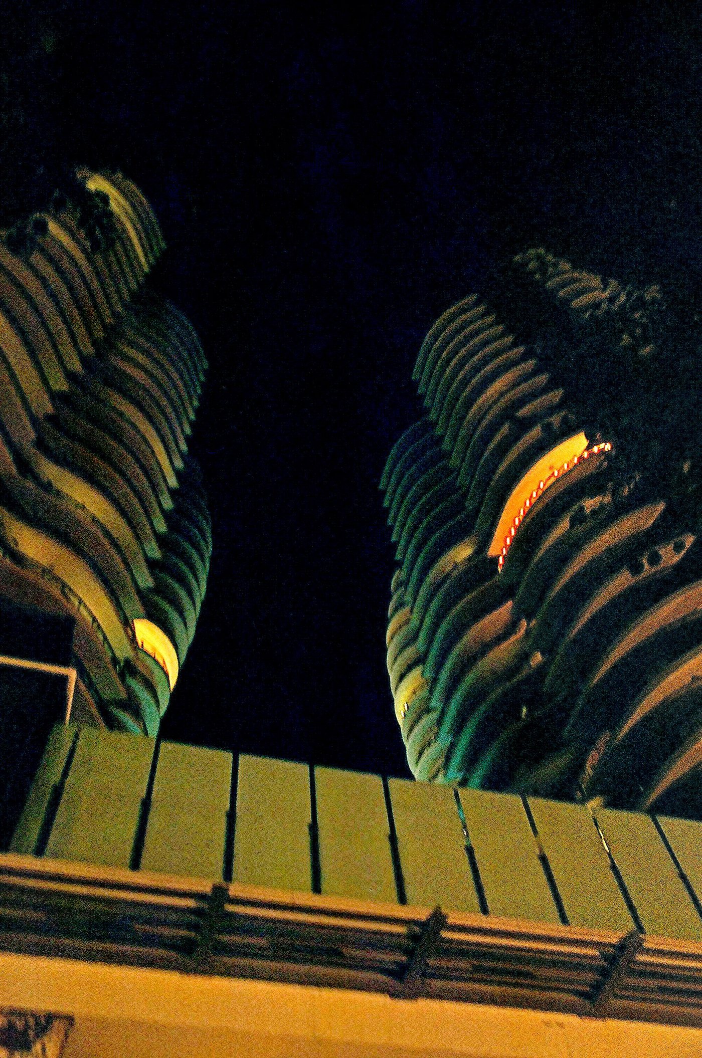 Looking up at night between two recently built residential towers with curved stacked balconies, their undersides lit orange against a black sky