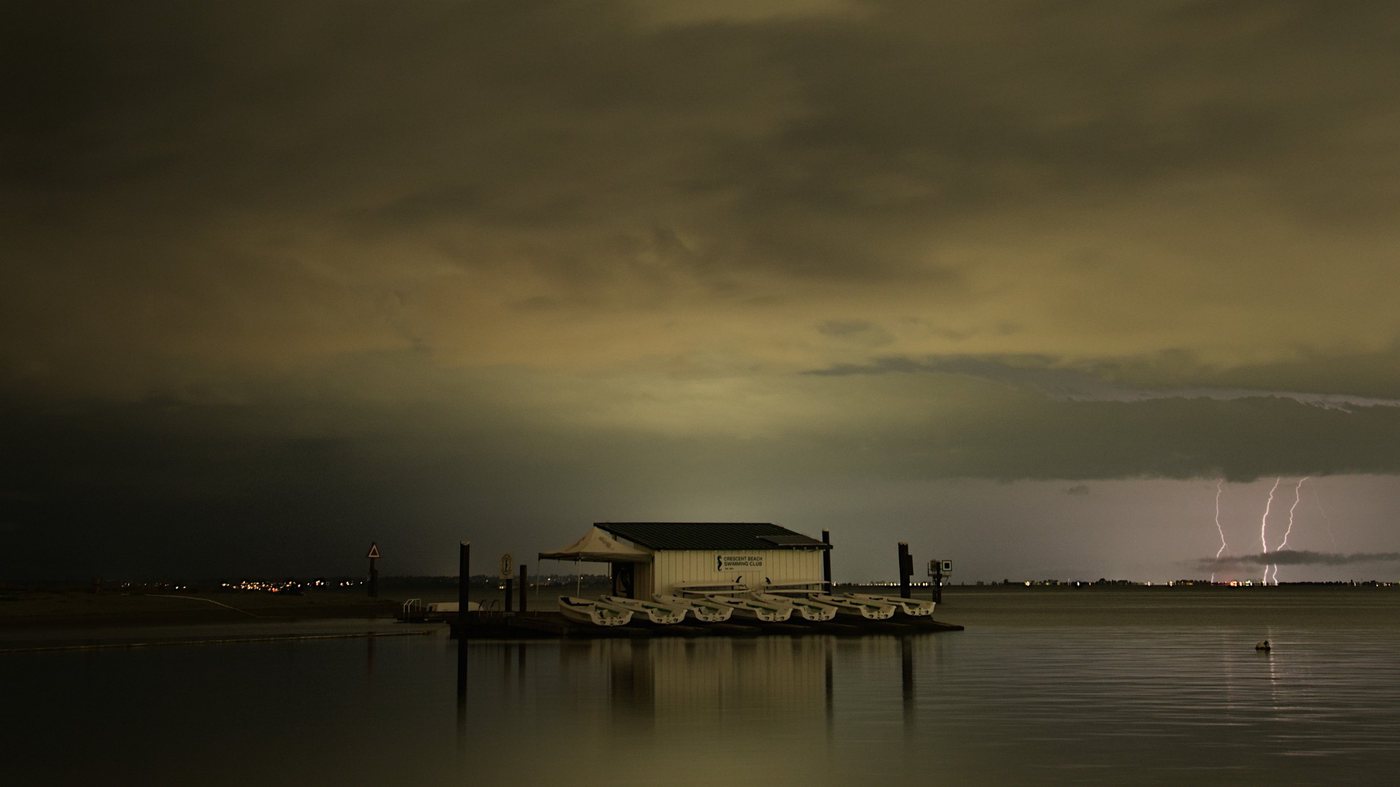 A floating dock of white rowboats beside the Crescent Beach Swimming Club hut on dark water, with two forks of lightning striking the horizon beneath a heavy sky