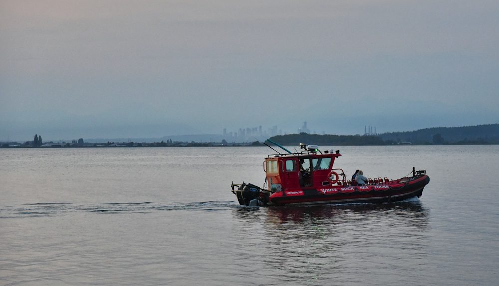 A small red tour boat crossing calm water at dusk, with a wooded spit in front of the distant tower cluster of Metrotown, hazy on the horizon