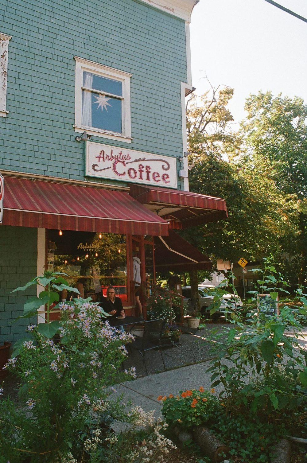 A green-shingled heritage corner building housing Arbutus Coffee, with a striped awning and a garden out front