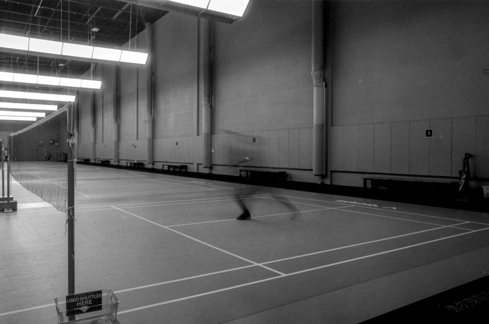 An empty sports hall with a single blurred figure mid-stride on a badminton court