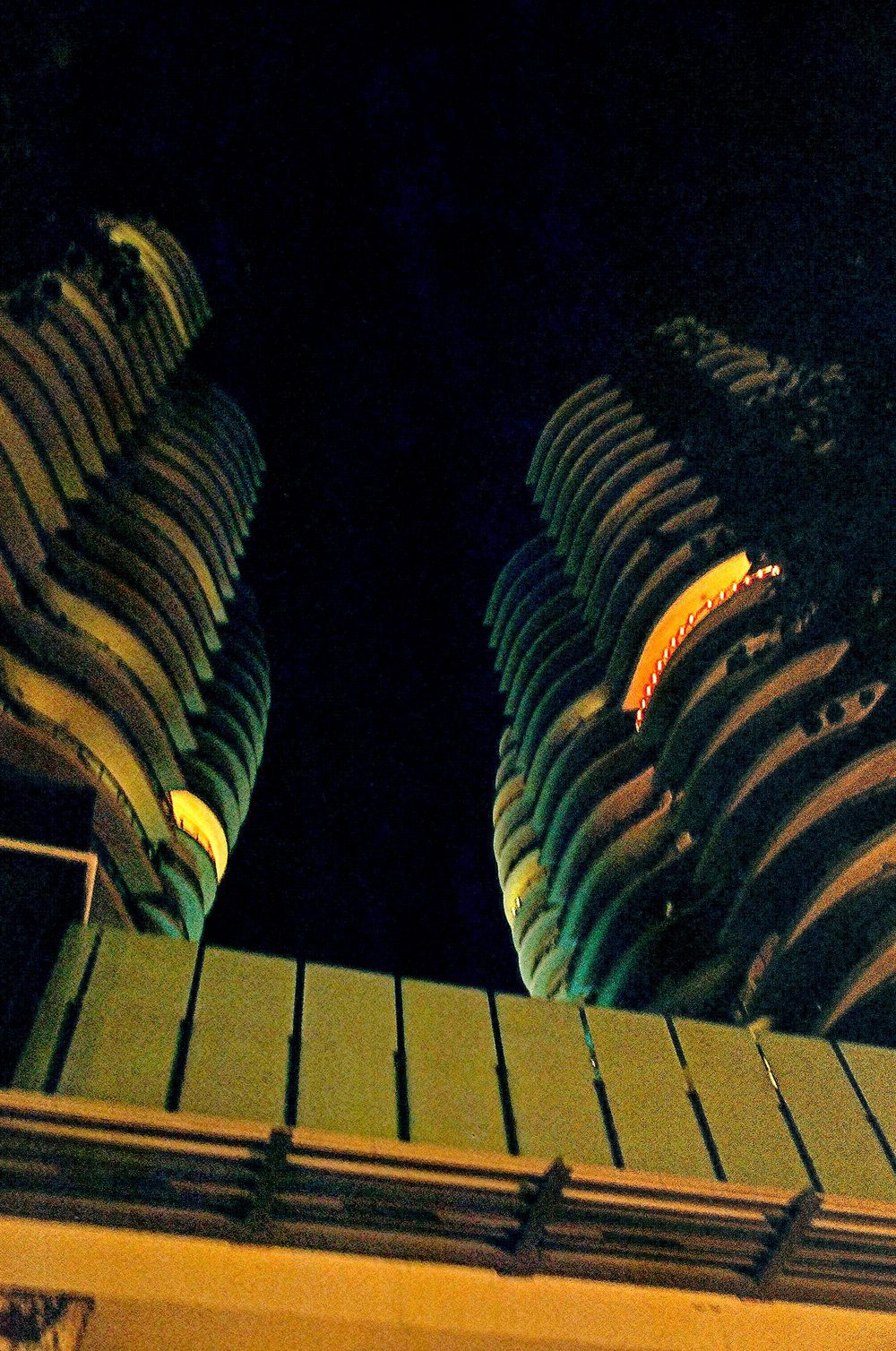 Looking up at night between two recently built residential towers with curved stacked balconies, their undersides lit orange against a black sky