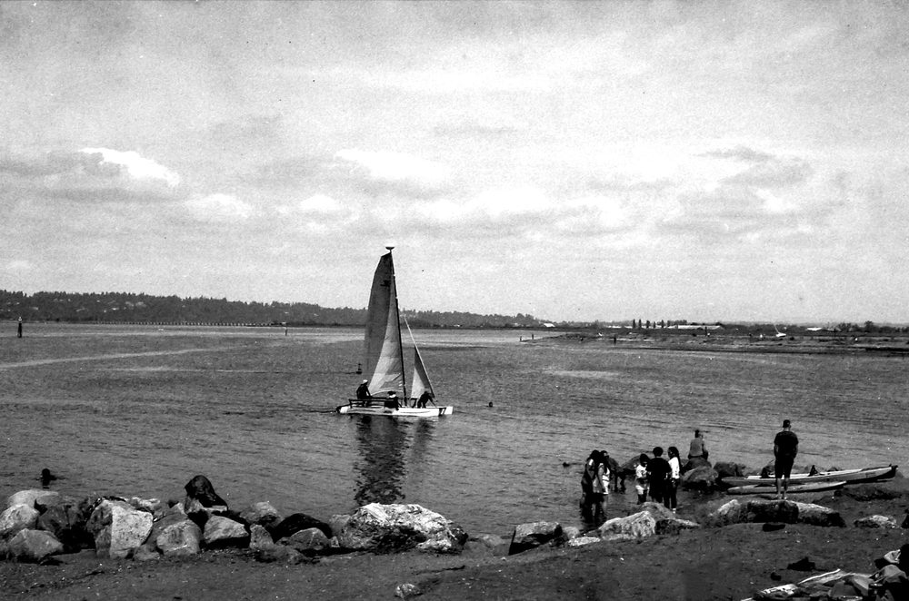 A small catamaran under sail close to shore, with people standing on the rocks watching