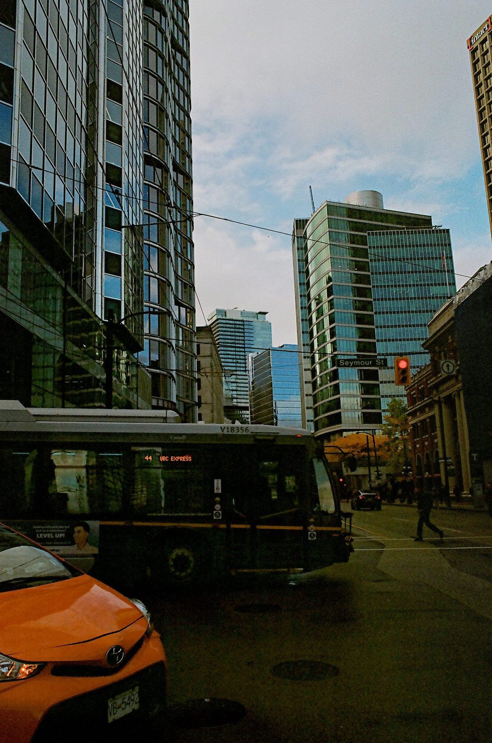 Downtown Vancouver glass office towers seen past a trolley bus at an intersection, an orange car in the foreground