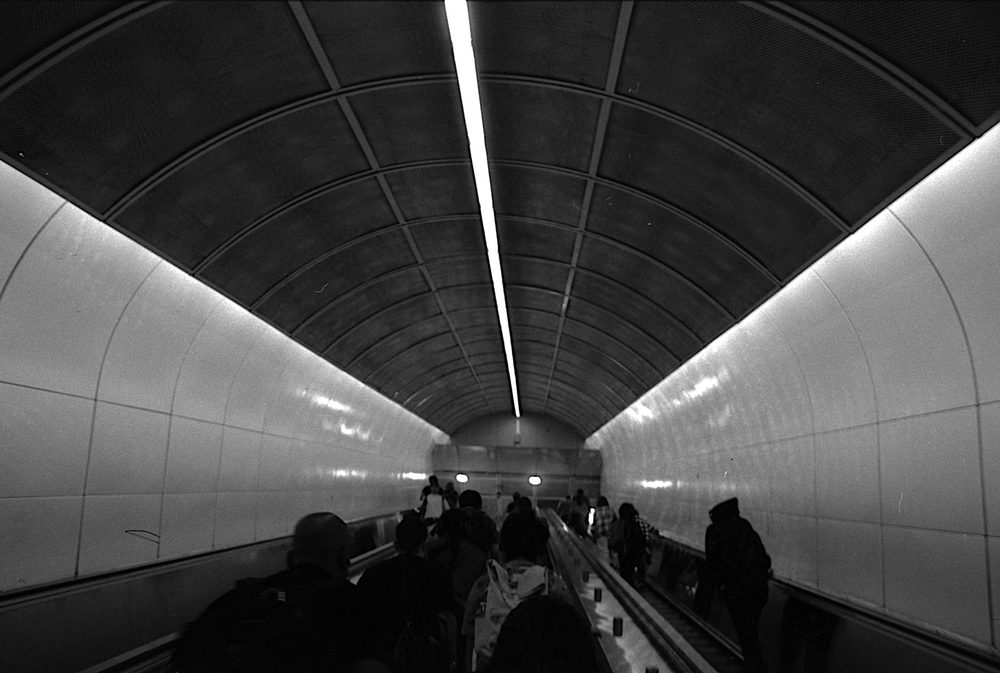 Passengers descending a long escalator through a tiled vaulted tunnel lit by a single strip light