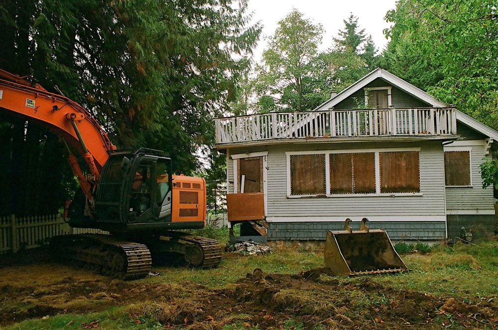 An orange excavator parked beside a boarded-up character house with a detached bucket on the lawn