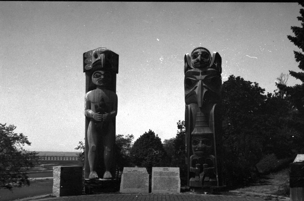 Two tall carved poles standing either side of a pair of stone interpretive plaques, with the sea and a long pier behind them