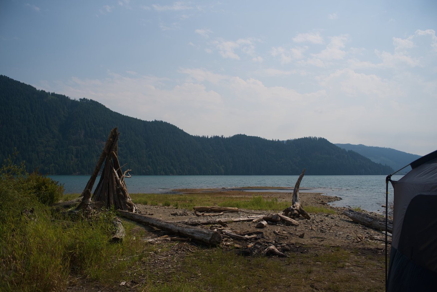 A lake ringed by forested hills, with bleached driftwood leaned together into a shelter on a gravel shore and the corner of a tent at the right